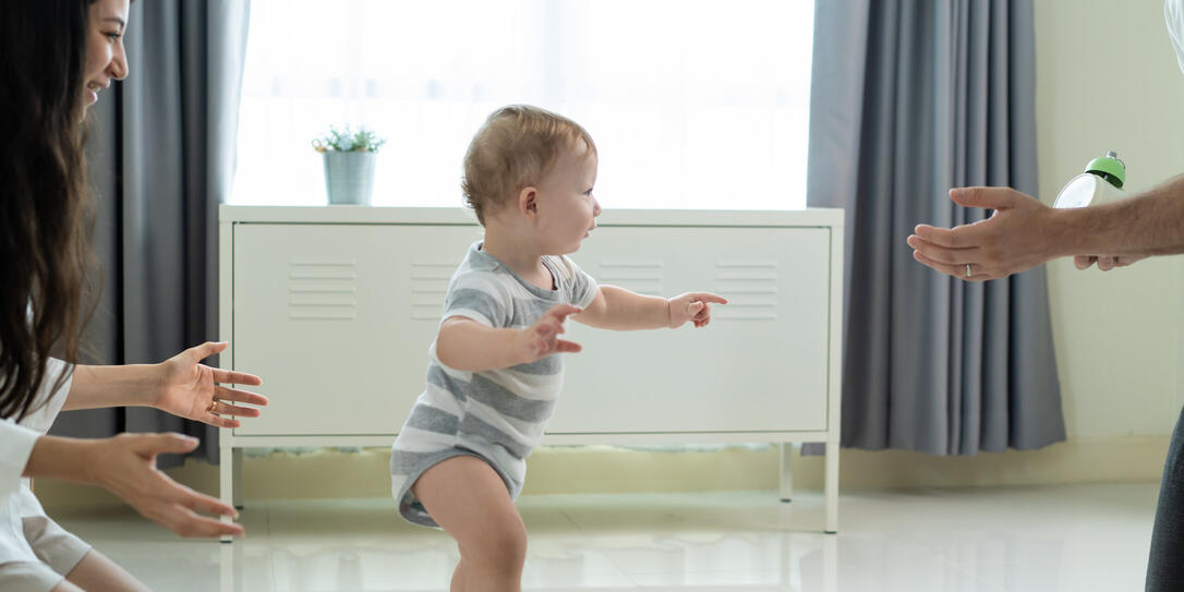 Caucasian young cute baby boy child learn to walk with parent in house. Happy family mother support little toddler son taking first step walk on floor to father to develop skill in living room at home