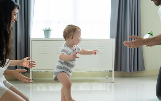 Caucasian young cute baby boy child learn to walk with parent in house. Happy family mother support little toddler son taking first step walk on floor to father to develop skill in living room at home