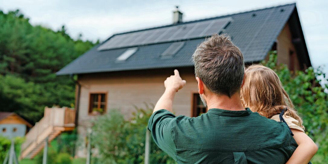 Rear view of dad holding her little girl in arms and showing at their house with installed solar panels. Alternative energy, saving resources and sustainable lifestyle concept.