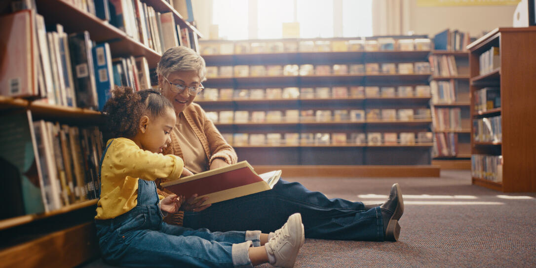 Library, learning and grandmother with grandchild on floor with book for reading, story and fun together. Education, child development and grandma with girl, storytelling and teaching while bonding